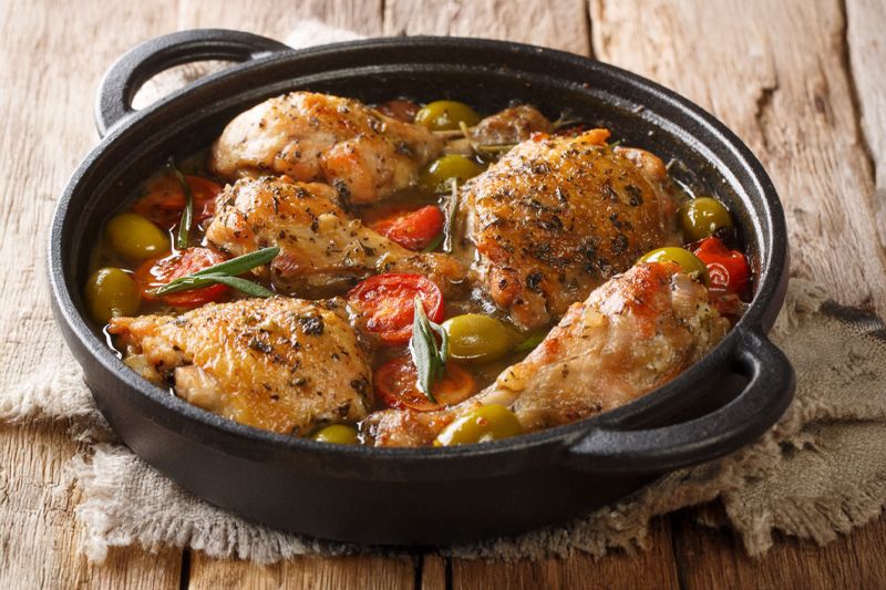 Cazuela de muslos de pollo con tomates cherry, aceitunas y cebolla dulce sobre una mesa de madera antigua