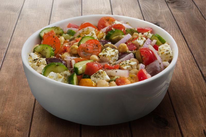Bol blanco lleno de ensalada de garbanzos, tomates cherry, cebolla dulce, pepino, pimiento, queso feta sobre una mesa de madera oscura