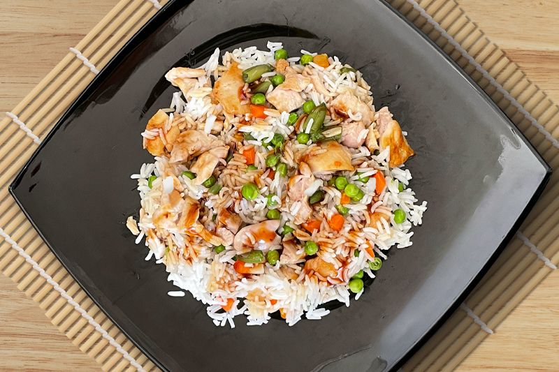 Ensalada de arroz con cebolla dulce, guisantes, judías verdes, zanahoria, pollo y salsa Teriyaki servida en un plato cuadrado negro.