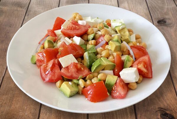 Plato blanco de ensalada de garbanzos, queso, tomate, aguacate y cebolla dulce sobre una mesa de madera marrón.