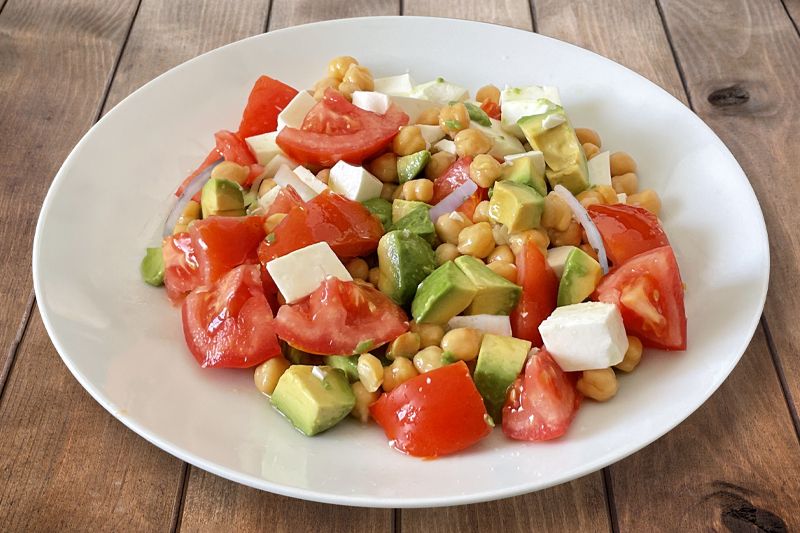 Plato blanco de ensalada de garbanzos, queso, tomate, aguacate y cebolla dulce sobre una mesa de madera marrón.