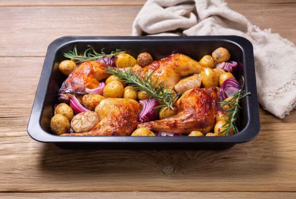 Pollo al horno con patatas pequeños, cebolla dulce en bandeja metálica negra sobre una mesa de madera