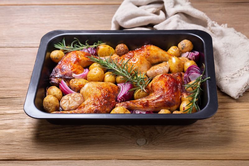 Pollo al horno con patatas pequeños, cebolla dulce en bandeja metálica negra sobre una mesa de madera