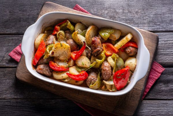 Fuente blanca sobre una tabla de madera de longanizas al horno con patatas, cebolla dulce y pimientos