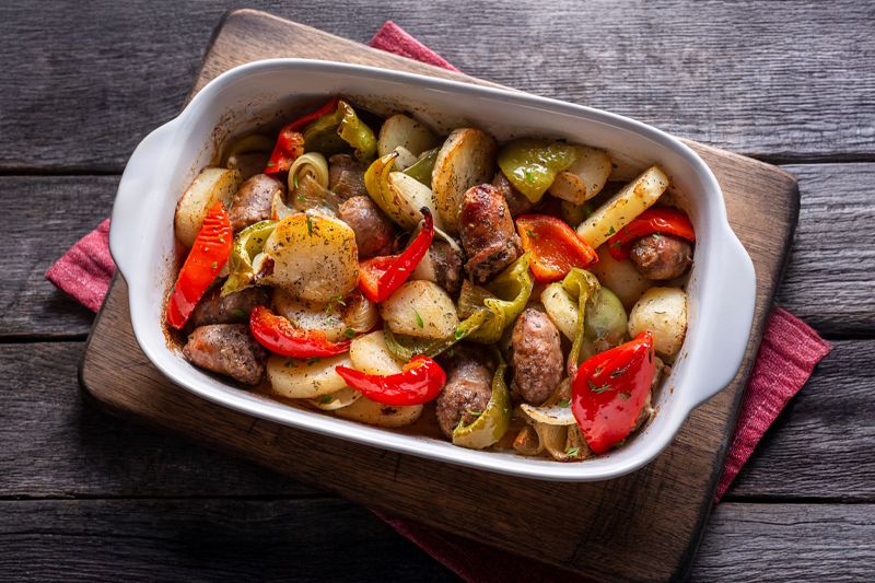 Fuente blanca sobre una tabla de madera de longanizas al horno con patatas, cebolla dulce y pimientos