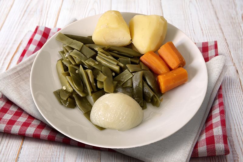 Plato de hervido valenciano compuesto por cebolla dulce, judías verdes, zanahoria y patata sobre unas servilletas en una mesa de madera blanca
