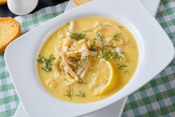 Plato blanco de sopa griega de limón con pollo sobre una mesa con mantel de cuadros verdes y blancos.