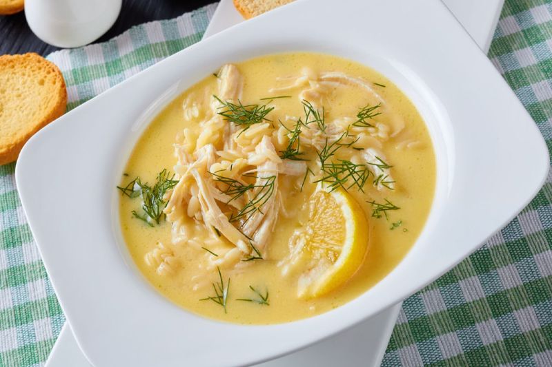 Plato blanco de sopa griega de limón con pollo sobre una mesa con mantel de cuadros verdes y blancos.