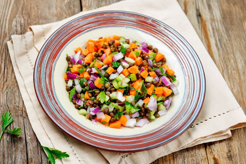 Plato de ensalada de lentejas, zanahorias, cebolla dulce y apio sobre una servilleta en una mesa de madera.