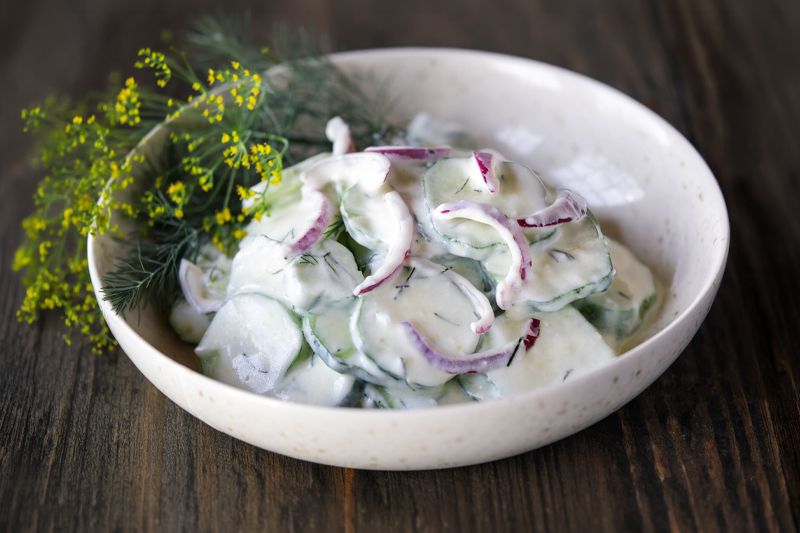 Cuenco blanco con ensalada cremosa de pepino y cebolla dulce o roja encurtida sobre una mesa de madera oscura.